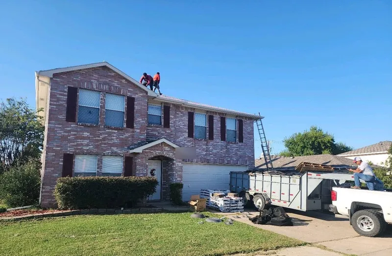 Roofers on the ridge of a two-story brick home, with shingle bundles and a dump trailer in the driveway